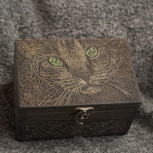 May include: A wooden box with a carved cat design. The cat has green eyes and a detailed face. The box has a hinged lid and a metal clasp.