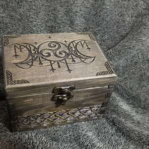 May include: A wooden box with a hinged lid and a decorative metal clasp. The top of the box is engraved with a triple moon symbol and Celtic knotwork. The box is stained a dark brown color.