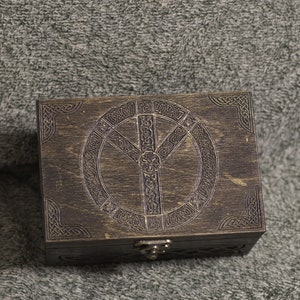 May include: A dark brown wooden box with a carved Celtic knot design and a metal latch. The box has a circular design with a Y-shaped symbol in the center.