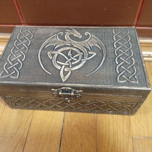 May include: A wooden box with a hinged lid, featuring a dragon and Celtic knot design. The box is stained in a dark brown color with silver accents. Intricate Celtic knot patterns are carved on the sides and top, and a metal clasp secures the front.