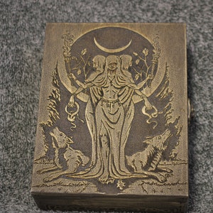 May include: A wooden box with a carved design of a triple goddess figure with a crescent moon above her head. The goddess is holding torches in each hand and is surrounded by trees and wolves. The box is painted in a gold and brown color.
