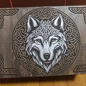 May include: A wooden box featuring a wolf design within a Celtic knot circle. The wolf has green eyes and a silver and white colour scheme. The box is stained dark brown, with Celtic knot corner accents and detailing.