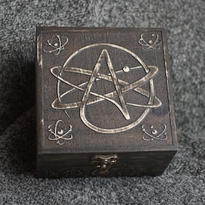 May include: A dark brown wooden box with a carved design of an anarchy symbol inside a circle. The box has a metal latch and four smaller carved designs of atoms on the sides.