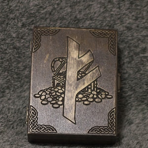 Könnte beinhalten: Eine rechteckige Holzbox mit einem geschnitzten Design. Die Box zeigt eine große Runen-Symbol und eine Schatztruhe mit Münzen. Aufwendige keltische Knotenmuster verzieren die Ecken. Die Box hat eine dunkle, verwitterte Oberfläche.