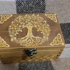 May include: A wooden box with a Celtic knot design and a tree of life carving on the lid. The box has a metal latch and is made of light brown wood.