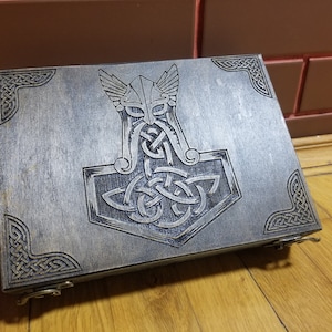 May include: A wooden box with a hinged lid, featuring an intricate carved design of a Viking helmet and hammer, with Celtic knotwork details. The box has a dark wood finish and silver hinges.