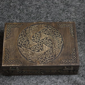 May include: A wooden box with a hinged lid, featuring an intricate Celtic knot design with two fish intertwined. The box is dark brown with a distressed finish.