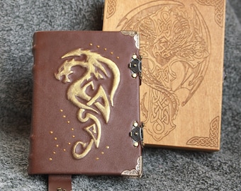 May include: Two leather-bound journals with gold dragon designs. The journal on the left has a brown cover with a gold dragon design and a gold clasp. The journal on the right has a light wood cover with a carved dragon design.