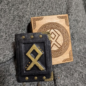 May include: A black leather journal with a gold rune symbol on the cover. The journal is attached to a chain with a tree charm. The journal is sitting on a gray fuzzy surface next to a wooden box with a similar rune symbol.
