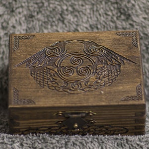May include: A dark brown wooden box with an engraved design on the lid. The design features two ravens, a Celtic knot, and spiral patterns. The box has a metal clasp and corner detailing. The box is made of wood.