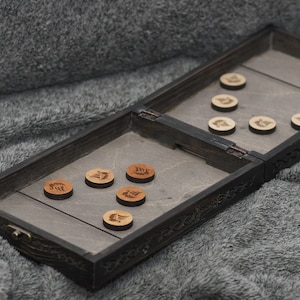 Pode incluir: Um tabuleiro de jogo de madeira com uma superfície de jogo cinza e uma moldura de madeira preta. O tabuleiro está aberto e mostra duas fileiras de peças de jogo de madeira com um design de navio. O tabuleiro está sobre uma superfície cinza e felpuda.