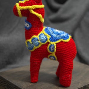 May include: A red crocheted Dala horse with yellow and blue details. The horse is standing on a wooden base.