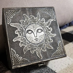 May include: A wooden box with a silver sun design on the lid. The sun has a face with closed eyes and a stylized design. The box is decorated with a Celtic knot pattern around the edges.