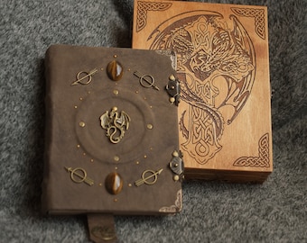 May include: Two leather-bound journals with decorative metal accents. One journal has a brown cover with a dragon design and the other has a wooden cover with a dragon and Celtic knot design.