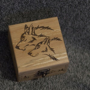 Könnte beinhalten: Eine Holzkiste mit einem Scharnierdeckel und einem Verschluss. Die Kiste ist mit einem Laser-gravierten Design von zwei Wolfsköpfen verziert.