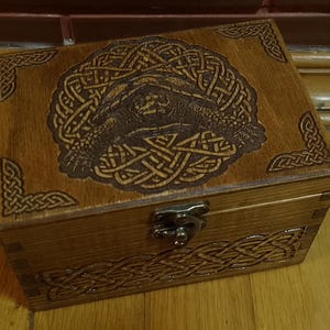 May include: A wooden box with a Celtic knot design and a metal latch. The box is made of dark wood and has a natural finish.