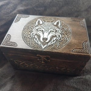 Può includere: Una scatola di legno con un design a testa di lupo intagliato sul coperchio. Il lupo è circondato da un motivo a nodo celtico. La scatola è di colore marrone scuro e ha un coperchio con cerniere.