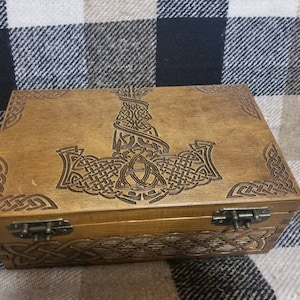 May include: A wooden box with a hinged lid, featuring an intricate Celtic knot design and a carved image of a hammer. The box has two brass hinges and a brown stain finish.