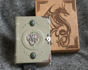 May include: A green leather journal with a silver dragon clasp and green gemstone accents. The journal is closed and sitting on a gray fuzzy surface. A wooden box with a carved dragon design is behind the journal.