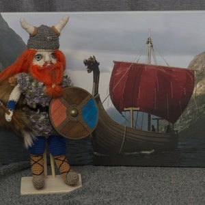 May include: Crocheted Viking warrior doll holding a shield and axe, standing on a wooden base. The doll is wearing a horned helmet, a red beard, and a blue tunic. The background is a painting of a Viking ship sailing on the water.