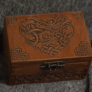 May include: A wooden box with a hinged lid and a carved Celtic knot design. The box is brown and has a brass latch.