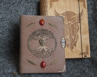 Pode incluir: Um diário de couro marrom com um design de árvore da vida e pedras preciosas vermelhas. O diário está fechado e tem um fecho de metal. Uma caixa de madeira com um design de coruja está atrás do diário.