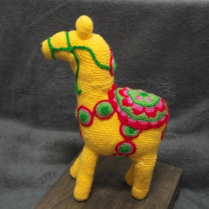 May include: A handmade, yellow crocheted horse figurine with green and red decorative accents. The horse has a detailed saddle and bridle, and is standing on a wooden surface. The figurine is approximately 20 cm tall.
