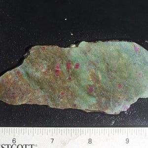 May include: A rough-cut stone with a mottled green and brown surface, speckled with small red inclusions. The stone is irregularly shaped and measures approximately 4 inches in length. A ruler is placed next to the stone for scale.
