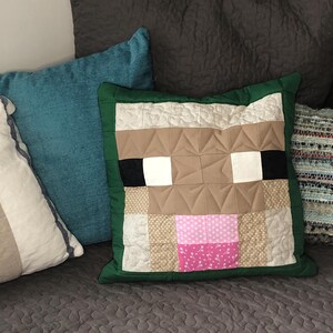 May include: A brown and white pillow with a green border, featuring a pixelated cow face design. The pillow is on a brown sofa with other pillows.