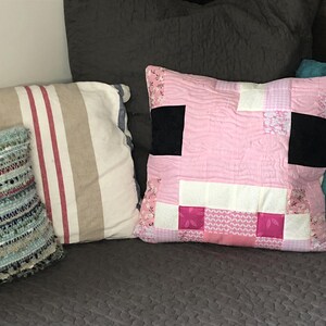 May include: A selection of decorative cushions in different styles. The main cushion is square and pink, with a patchwork design that looks like a pig's face, with black squares for eyes and white and pink blocks for the snout. Other cushions have striped and textured patterns.