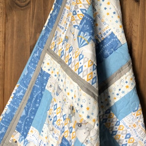May include: A patchwork quilt with a circus theme. The quilt features blue, white, grey, and yellow fabrics with circus-related imagery, including elephants, stars, and text. The quilt is trimmed with a grey border and has a quilted pattern.