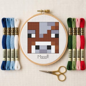 May include: A cross-stitch project in a wooden hoop featuring a pixelated brown and white cow face with the text Mooo!!!, flanked by four blue and white embroidery floss skeins on the left and four green, pink, and red skeins on the right.