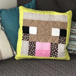 以下が含まれることがあります： 黄色の枕に、茶色、白、ピンクのMinecraft風の羊の顔のデザインが施されています。枕はパッチワーク生地で作られており、黄色の縁取りがあります。