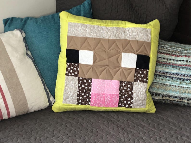 以下が含まれることがあります： 黄色の枕に、茶色、白、ピンクのMinecraft風の羊の顔のデザインが施されています。枕はパッチワーク生地で作られており、黄色の縁取りがあります。