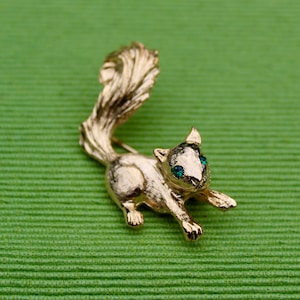 May include: A gold-toned squirrel brooch with green gem eyes. The squirrel is depicted with its tail curled up and its head turned to the side.