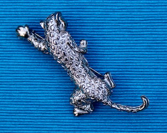 May include: Silver-toned brooch of a leaping leopard with a spotted pattern. The leopard is holding a ball in its paw.