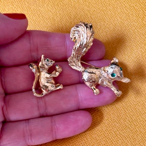 May include: Two gold-toned squirrel brooches. One is a seated squirrel, the other is a running squirrel. Both have textured fur and green gemstone eyes. The brooches are held in a hand against a yellow background.