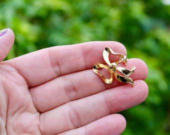 May include: A gold-toned bow brooch with a detailed design. The brooch is held in a hand against a green background.