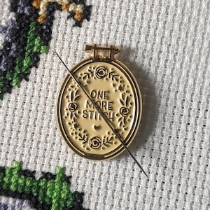 May include: A gold-coloured metal embroidery hoop with the text "ONE MORE STITCH" inside. The hoop is on a white Aida cloth with a green and grey cross-stitch design.