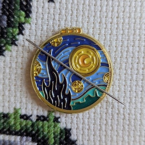 May include: A circular enamel pin with a starry night design and a gold edge. The pin shows a swirling blue sky, a crescent moon, and a stylized landscape. A silver needle is placed across the pin.