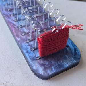 May include: A clear acrylic yarn holder with a marbled blue, purple, and white base. The holder has multiple slots to hold yarn and a bundle of red yarn is attached. The yarn holder is on a white surface.