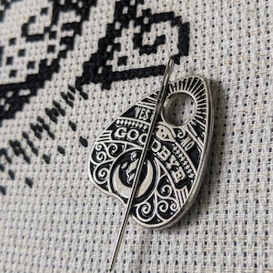 May include: A silver-coloured Ouija board planchette with black detailing and the words "Yes", "No", "Goodbye", and "Good Bye" on it. The planchette is on a white and black cross-stitch fabric.