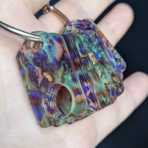 May include: A multi-coloured, rectangular keyring with a silver ring. The keyring has a marbled effect with shades of blue, green, and purple. It features two circular holes and a textured surface. The keyring is held in a hand.