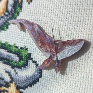 May include: A cross-stitch project in a wooden hoop shows a dragon design and a whale-shaped pin. The whale pin is a mix of purple, red, and white, with a needle inserted through it and the fabric. The hoop is approximately 15 cm in diameter.