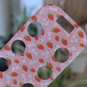 May include: A pink phone case decorated with a pattern of red strawberries and white flowers. The case features circular cutouts for the camera, buttons, and charging port. The case is made of a smooth, glossy material.