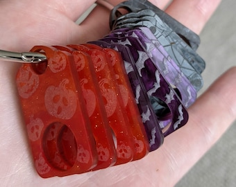 Puede incluir: Un juego de cinco llaveros con un anillo plateado. Los llaveros están hechos de plástico transparente con diferentes diseños de temática de Halloween. Un llavero es rojo con un diseño de calabaza, y los demás son morados y grises con diseños de murciélagos y telarañas.