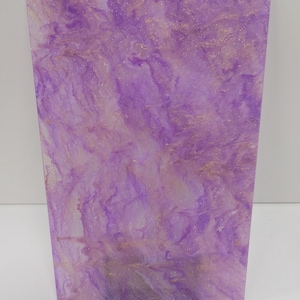 May include: An abstract art piece with a flowing design in shades of purple and gold. The artwork has a marbled effect, with the colours blending together. The piece is displayed on a black stand. The dimensions are not visible.