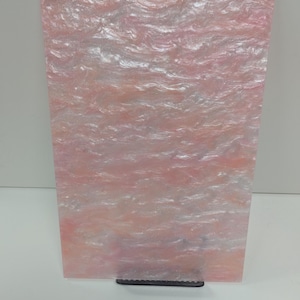 May include: A rectangular acrylic sheet with a pink and white swirling pattern. The material has an iridescent sheen, creating a wave-like texture. The sheet is displayed vertically, resting on a black support.