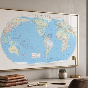 Peut inclure: Une carte murale rectangulaire intitulée THE WORLD présentant un fond océanique bleu clair et des masses terrestres multicolores. La carte est encadrée par un fin cadre doré et affiche divers pays, continents et étiquettes géographiques.