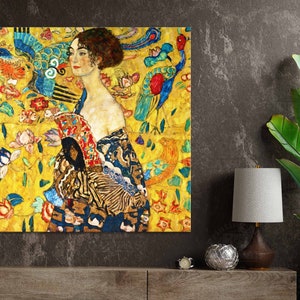 May include: A framed print of a Gustav Klimt painting. The artwork features a woman with brown hair and a red lip, wearing a patterned robe. The background is a vibrant yellow with floral and bird motifs, with accents of blue, red, and green.